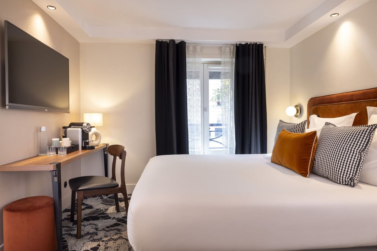 A hotel room with a bed, a desk with a chair, a television, and a window with curtains.