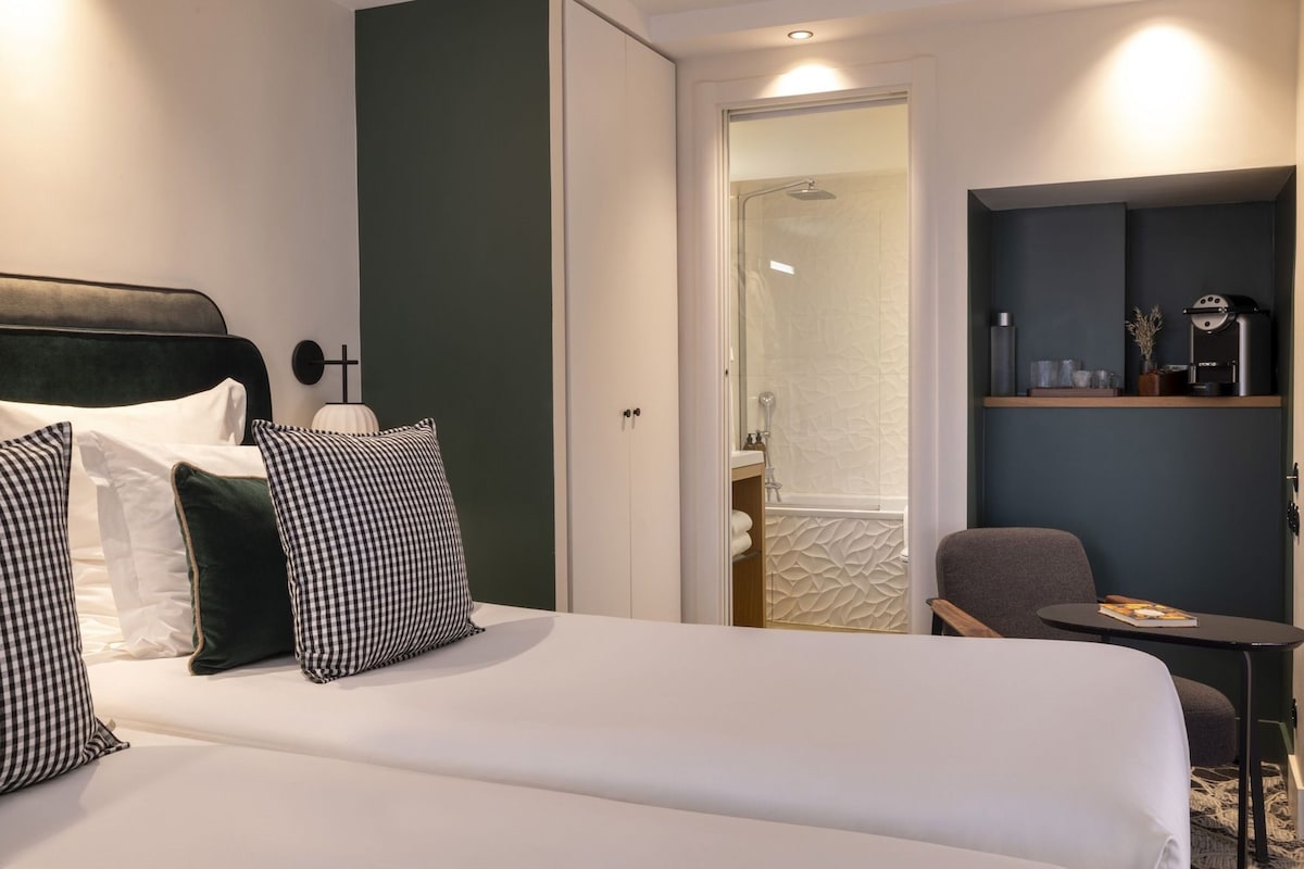 A hotel room with a bed, a chair, a small table, a coffee machine, and a shower area.