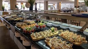 Un bufé con diversos alimentos y bebidas, que incluye ensaladas, sándwiches y postres, exhibidos sobre un mostrador con vitrina de vidrio.
