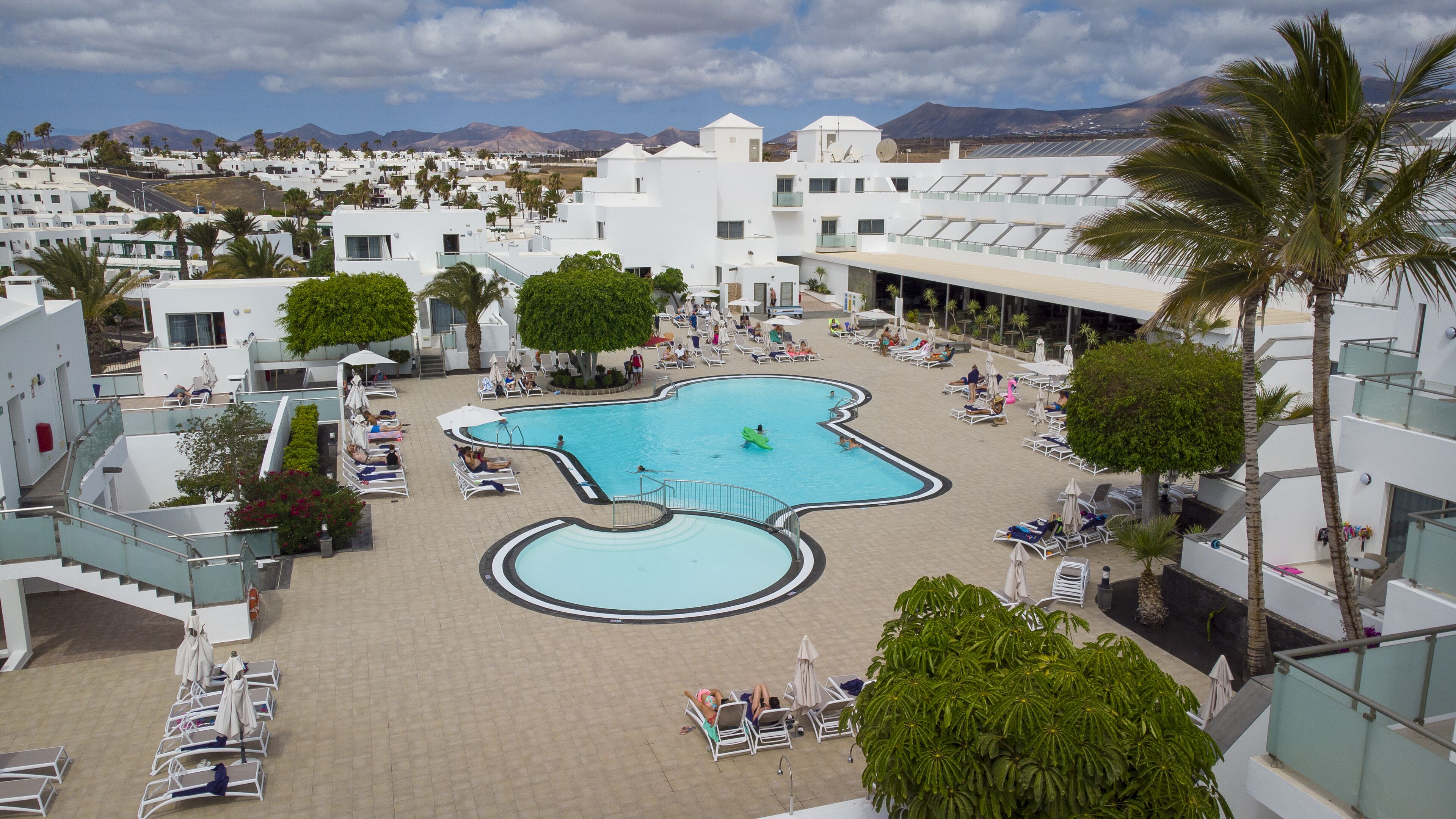 A resort with a central swimming pool, sun loungers, and palm trees.