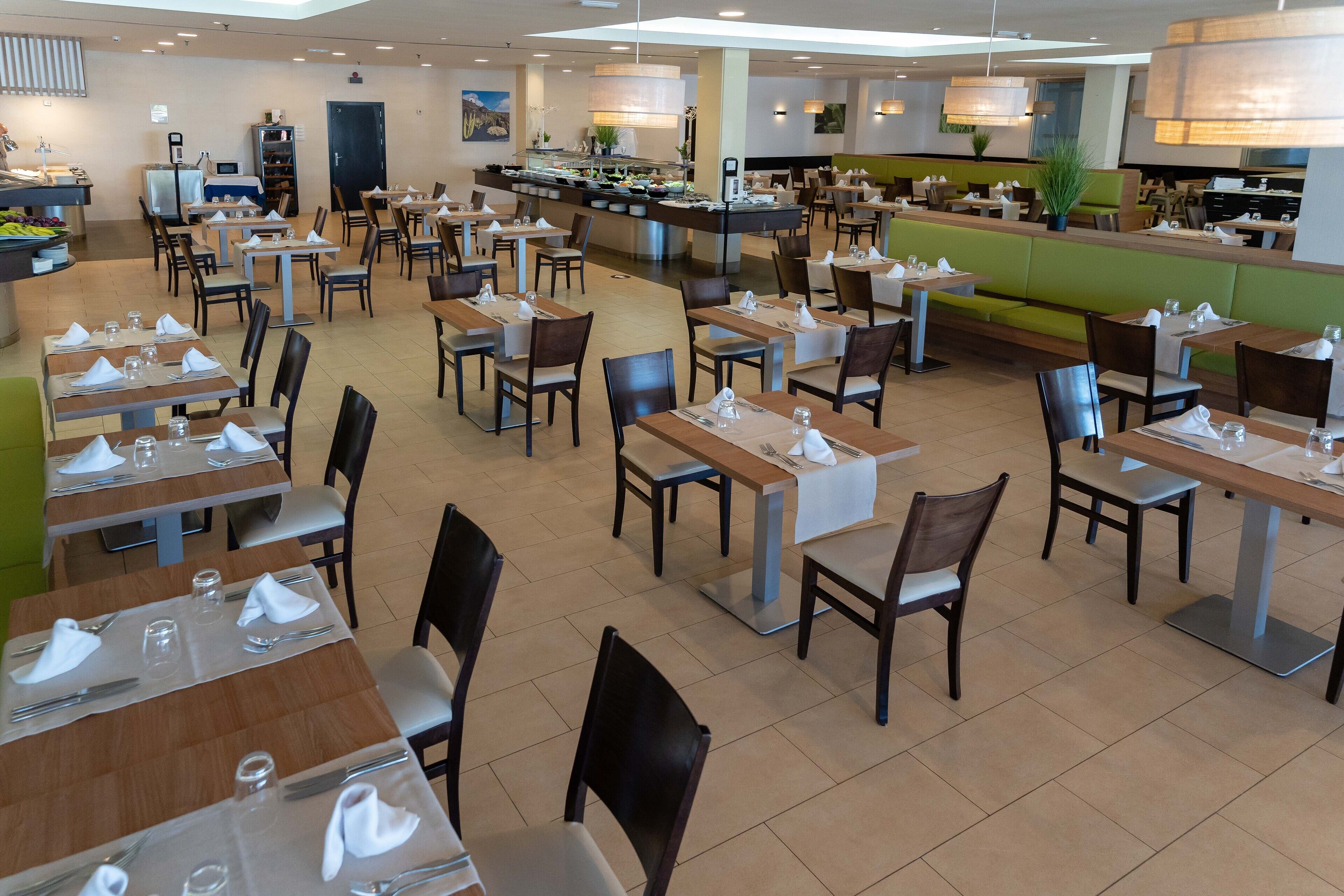 Uma ampla área de refeições com várias mesas e cadeiras, um balcão de buffet e uma cozinha ao fundo.