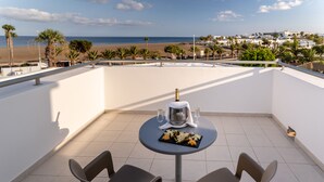 Una terraza con una mesa dispuesta para dos personas, una botella de champán y un plato de aperitivos. La terraza tiene vista a una playa y palmeras.