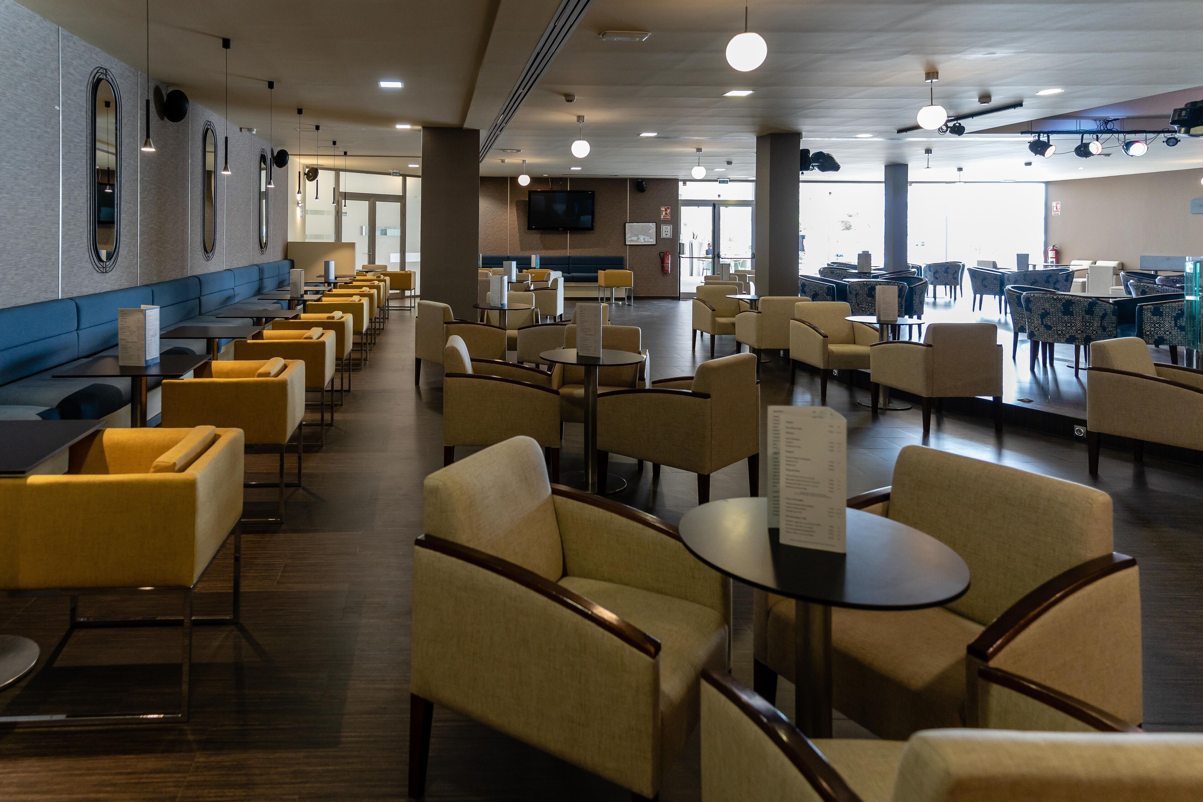 A spacious indoor seating area with multiple tables and chairs, a flat-screen TV mounted on the wall, and a blue cushioned bench along the wall.