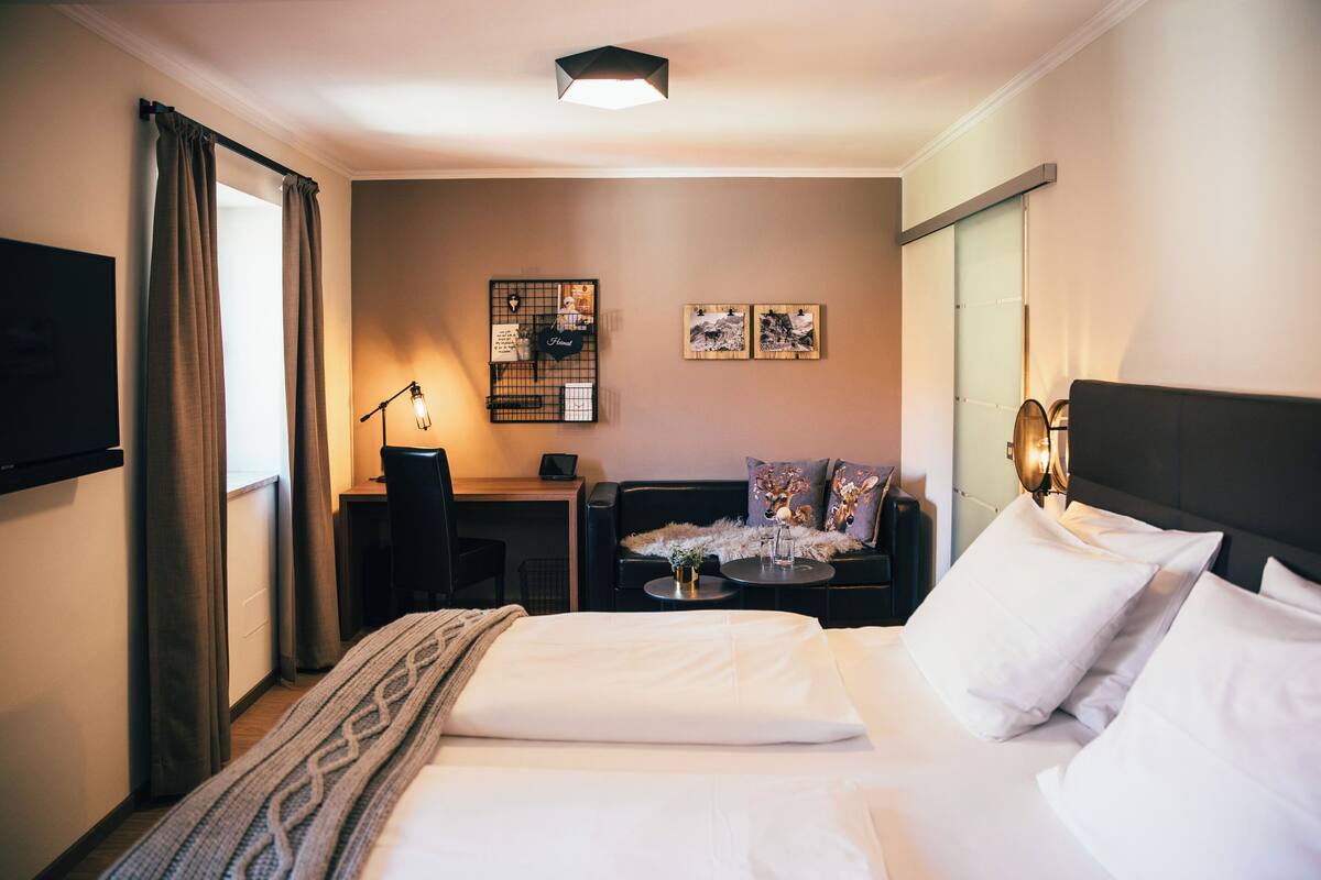 A hotel room with a bed, a desk, a chair, and a small table. There are curtains, a lamp, and wall art.