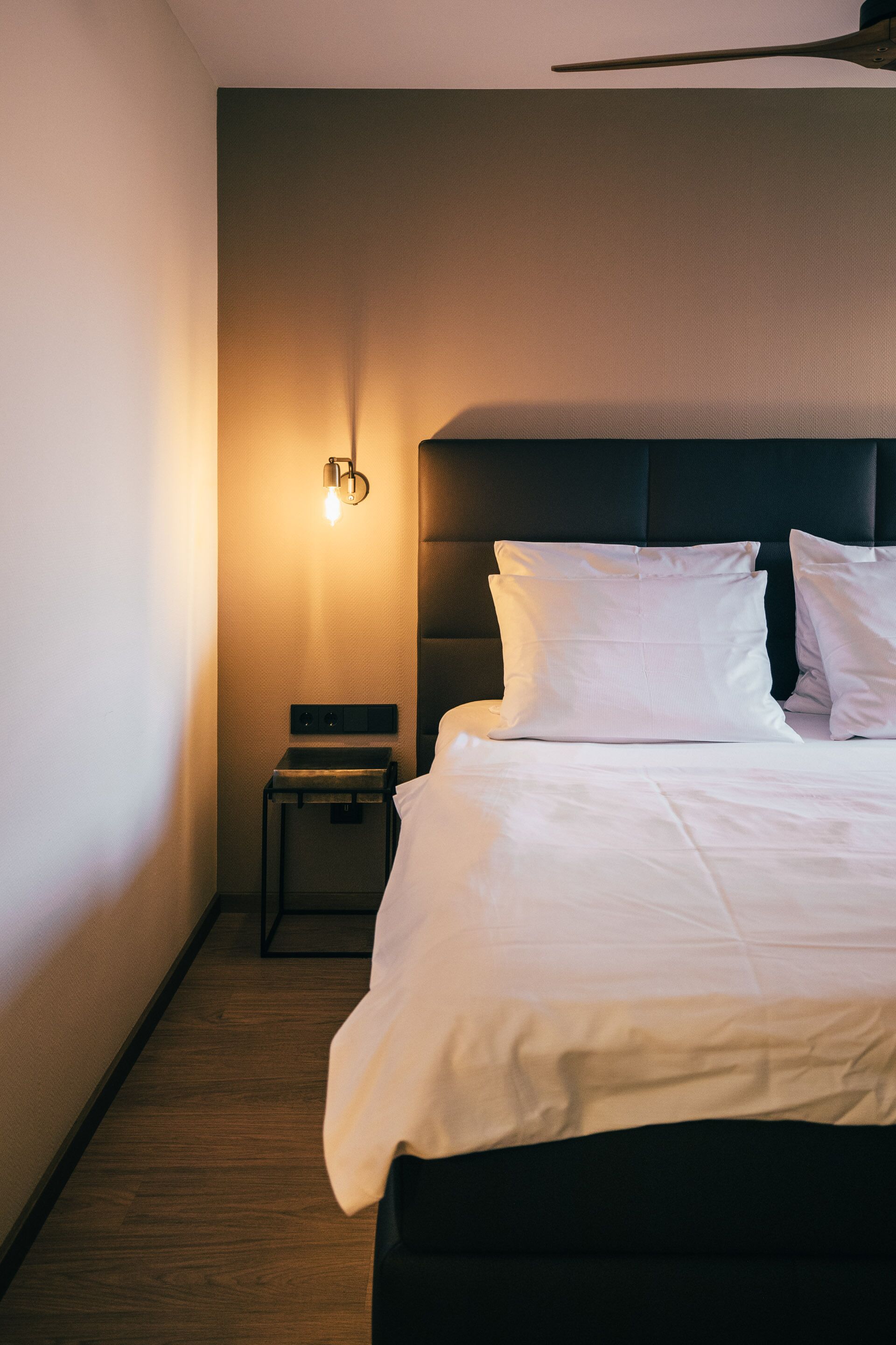 Siisti hotellihuone, jossa on huolitellusti pedattu sänky, yöpöytä ja seinään kiinnitetty valaisin.