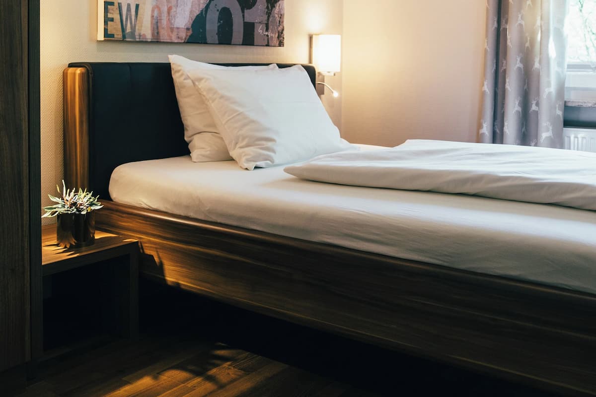 A hotel room with a bed, a nightstand, a wall-mounted lamp, a window with curtains, and a large poster on the wall.