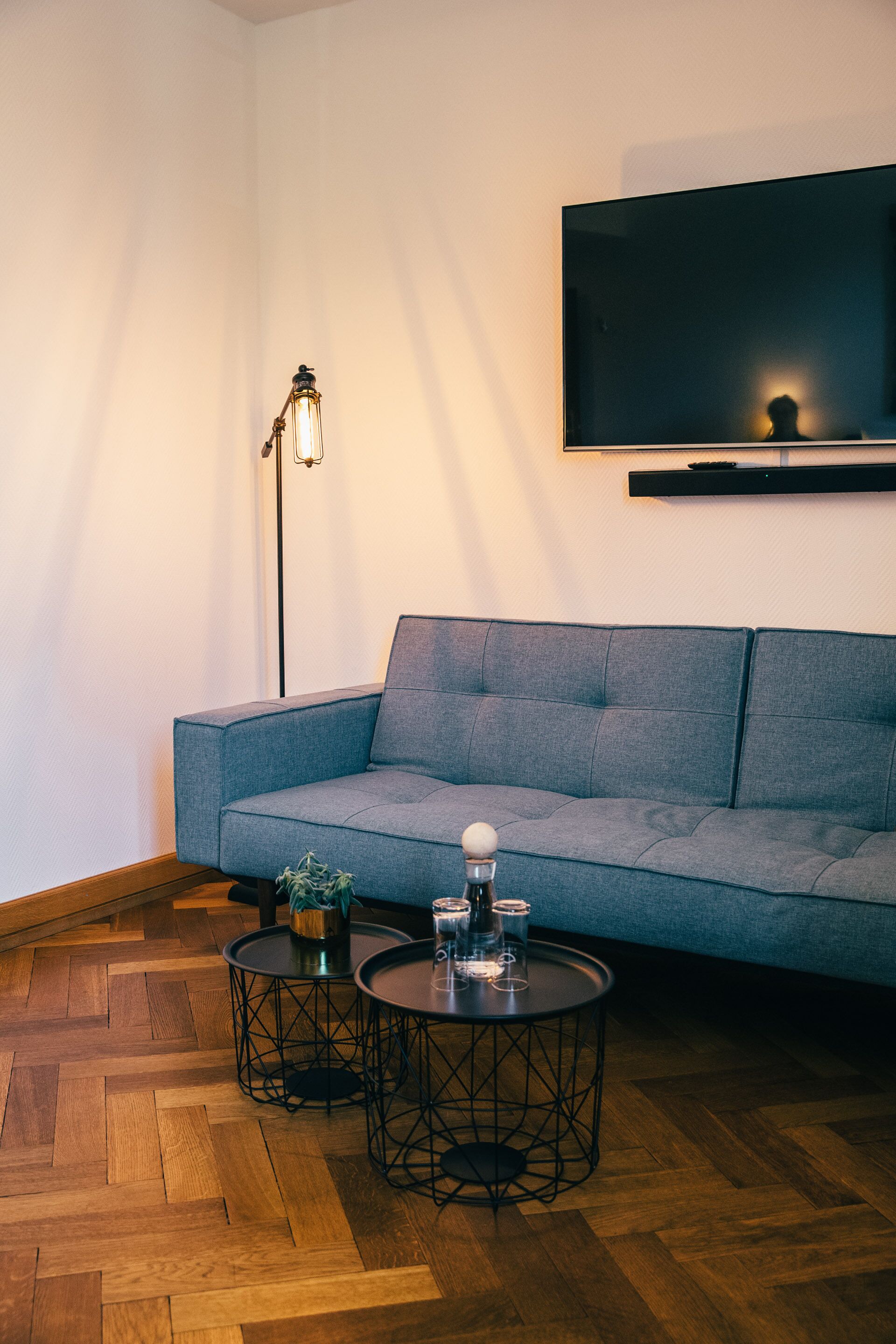 Una sala de estar moderna con un sofá gris, dos mesas redondas decoradas, una lámpara de pie y un televisor montado en la pared.