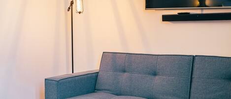 Una sala de estar moderna con un sofá gris, dos mesas redondas decoradas, una lámpara de pie y un televisor montado en la pared.