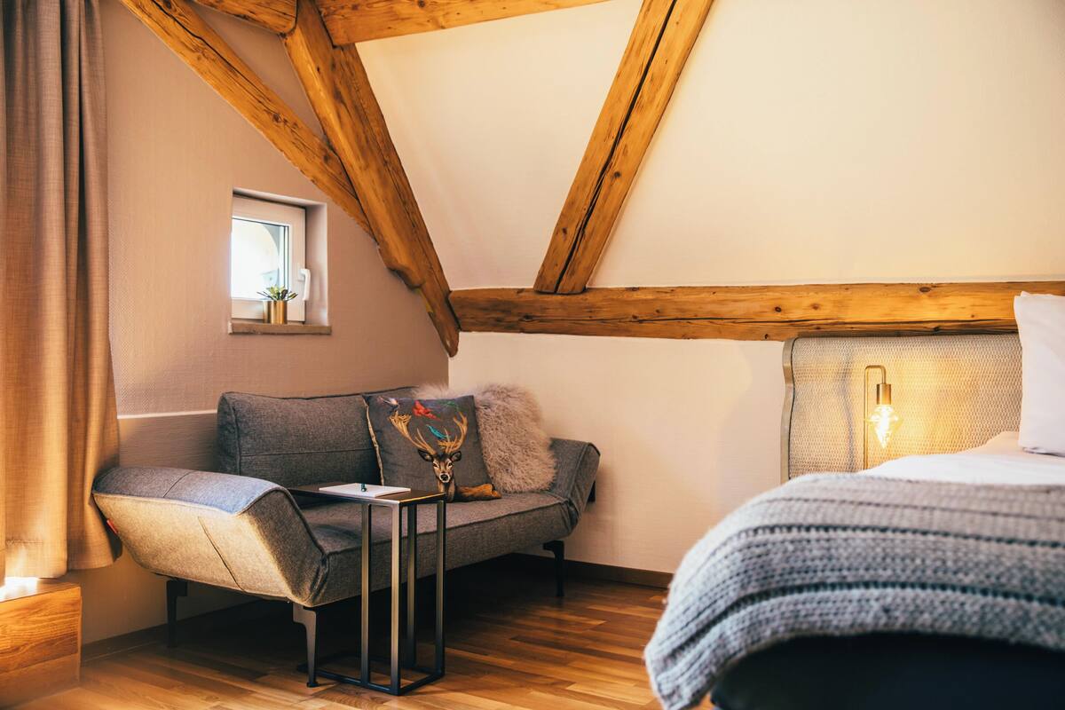 A cozy room with a wooden ceiling, a gray sofa, a small table, and a bed with a white headboard.