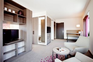 Une chambre d’hôtel moderne dotée d’une télévision à écran plat, d’un coin salon avec une table ronde et d’un dressing.