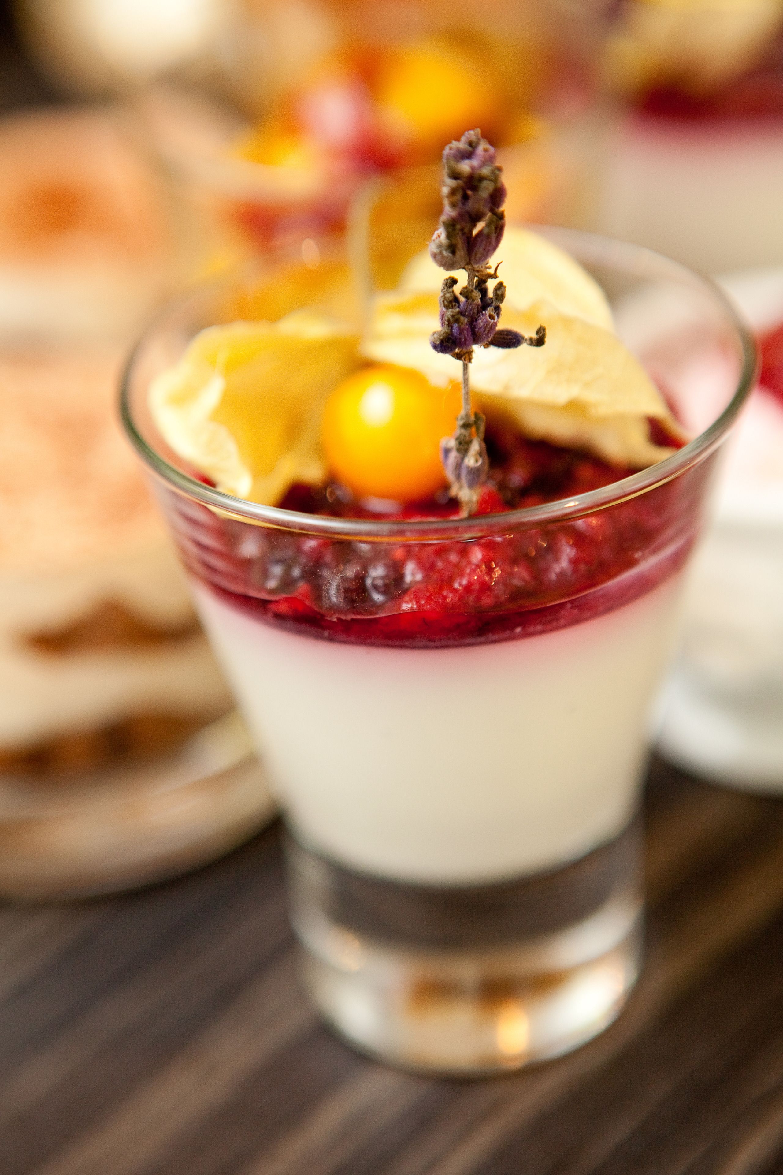A dessert in a clear glass with layers of cream and berries, garnished with a yellow fruit and a sprig of lavender.