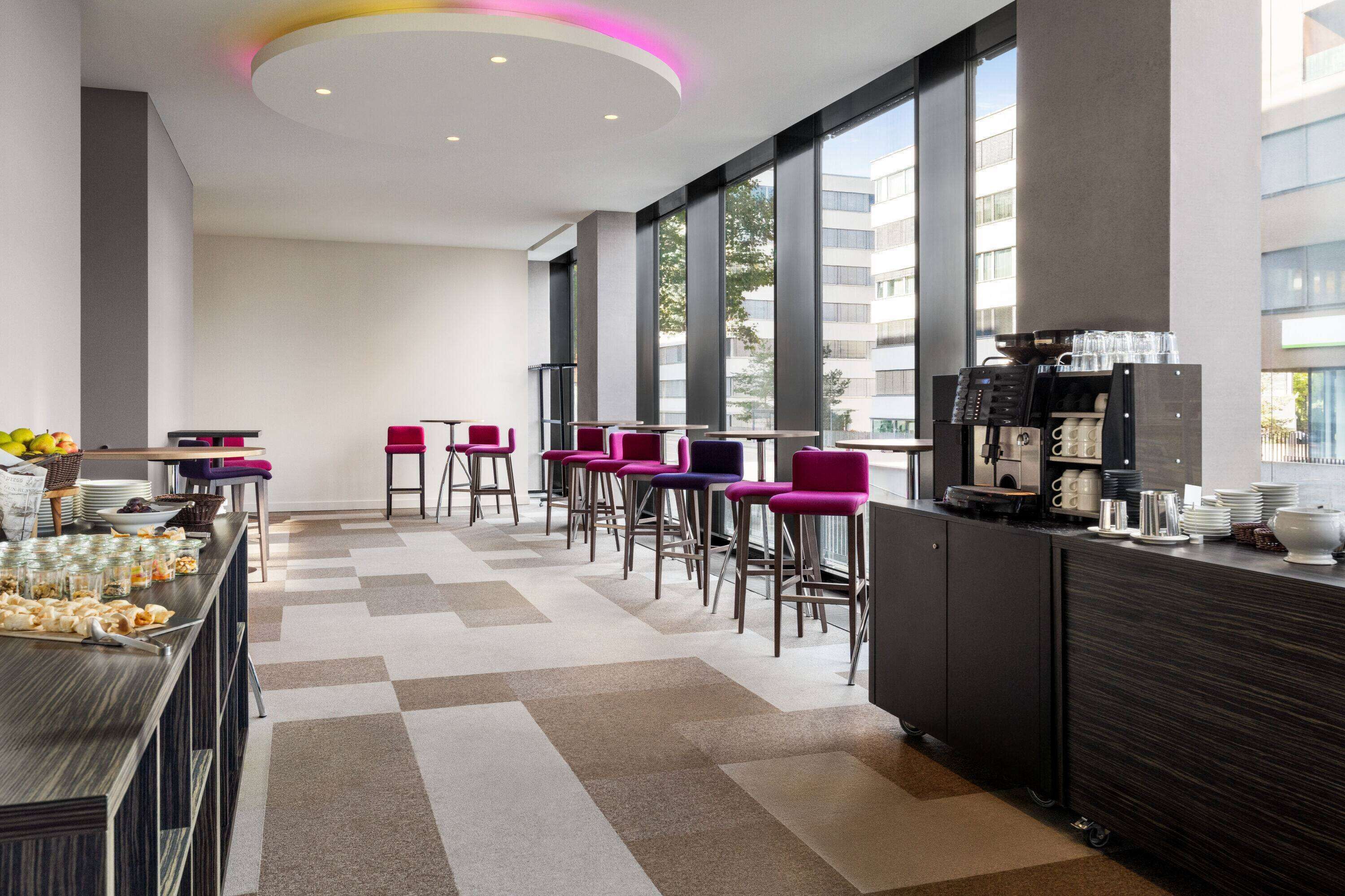 Un moderno hall de hotel con mostrador de recepción, zona de bar y asientos con sillas moradas.