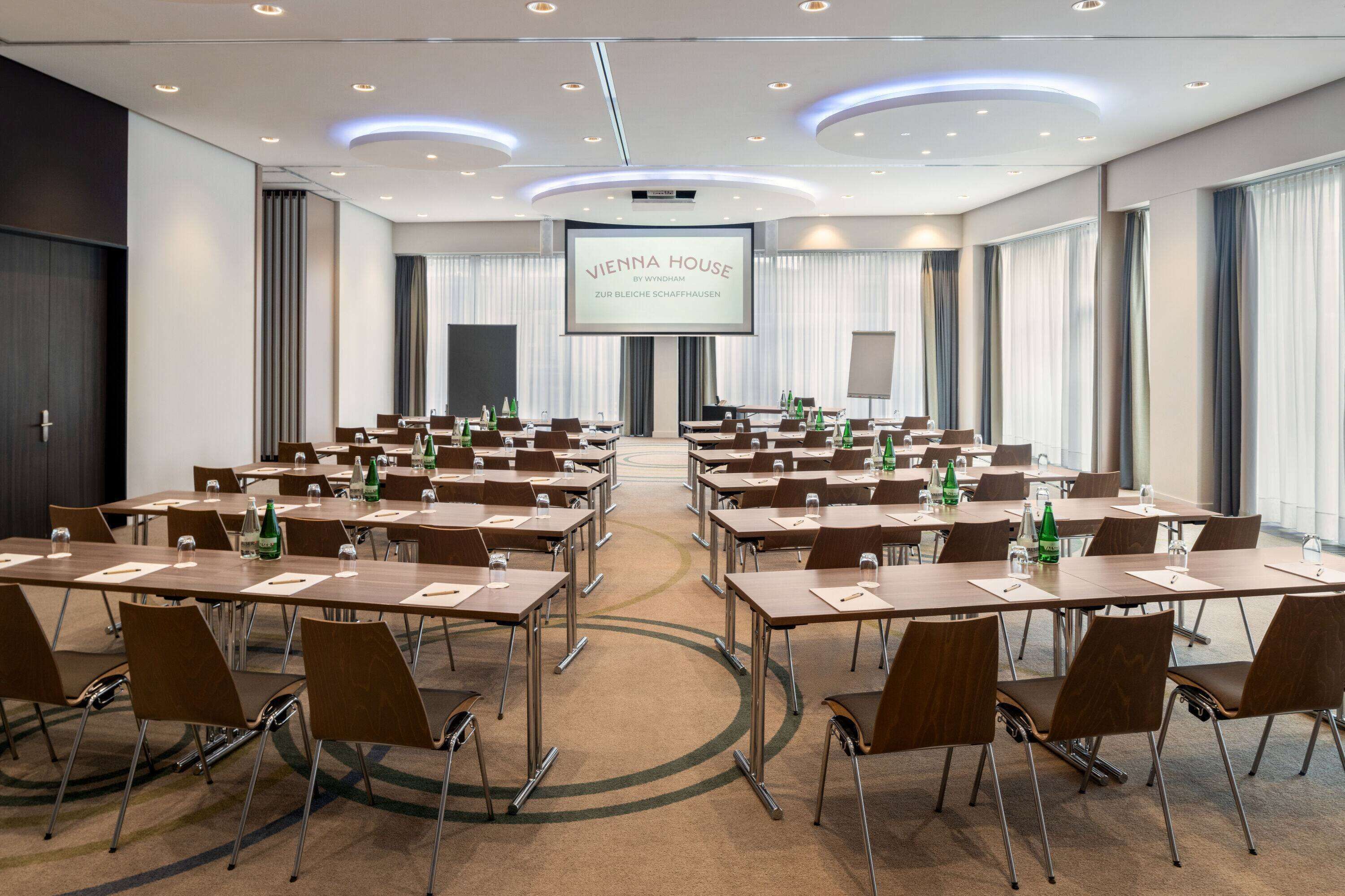 A conference room with multiple tables and chairs, a large screen at the front, and a carpeted floor.