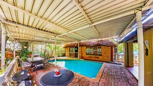 Un espace extérieur avec piscine, comprenant un patio couvert, une piscine en briques et un bâtiment doté d’un toit de chaume.