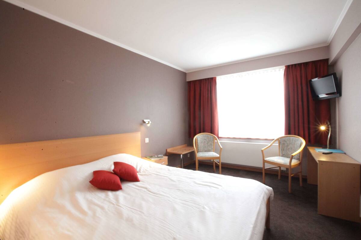 Ein Hotelzimmer mit einem großen Bett, zwei Sesseln, einem Schreibtisch, einem Fernseher und einem Fenster mit Vorhängen.