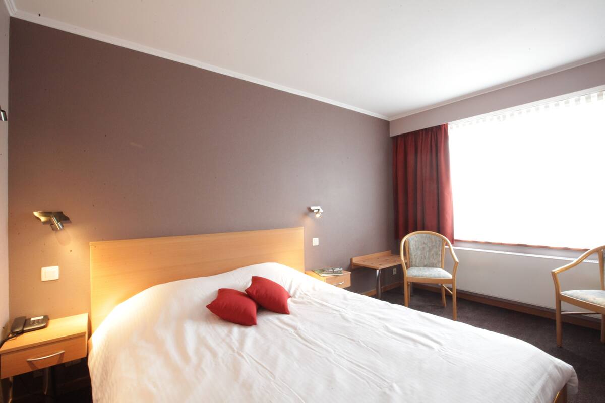 Ein Hotelzimmer mit einem großen Bett, zwei roten Kissen, einem hölzernen Kopfteil, einem Schreibtisch mit Stuhl und einem Fenster mit Vorhängen.