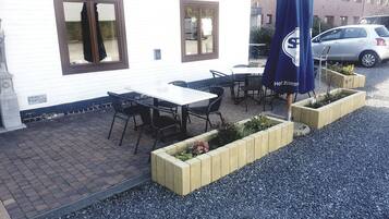 Een terras met tafels en stoelen, een blauwe parasol, en plantenbakken met planten.
