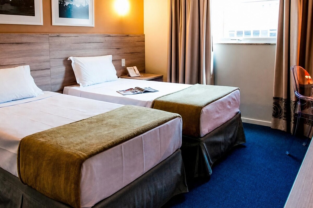 Habitación de hotel con dos camas, cabecero de madera, mesita de noche con teléfono y ventana con cortinas.