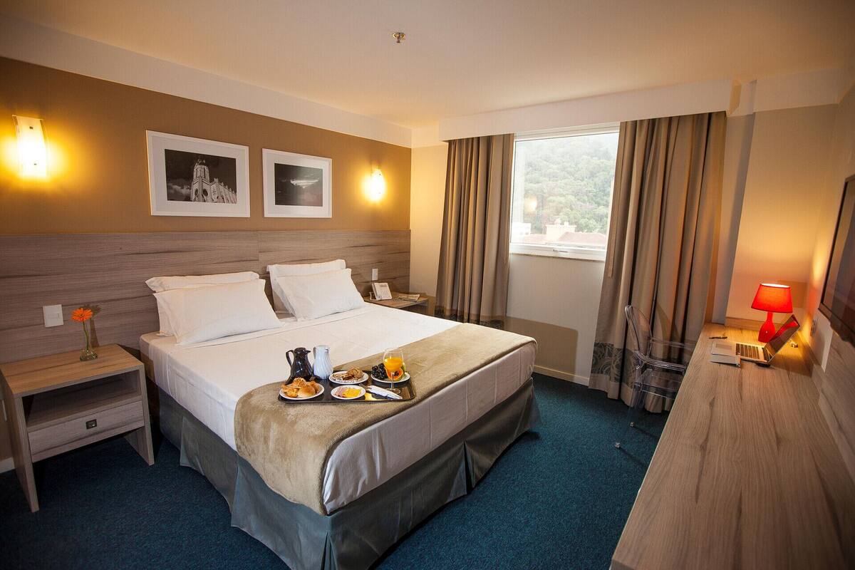 Una habitación de hotel con cama, mesitas de noche, escritorio, lámpara y ventana con cortinas.