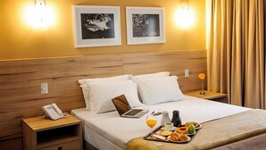 A hotel room with a wooden headboard, two framed pictures on the wall, a bedside table with a phone, a laptop, and a tray with breakfast items. - Intercity Teresopolis (Teresópolis)
