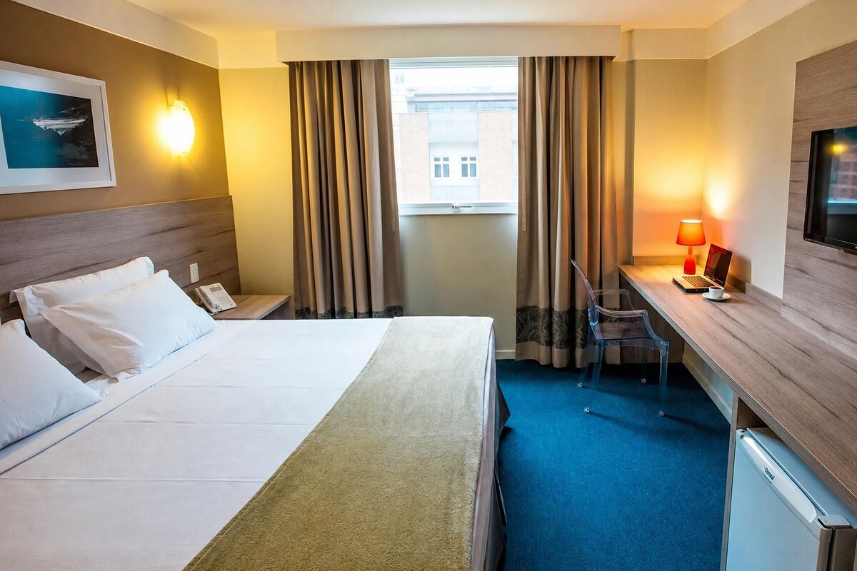 Habitación de hotel con cama, escritorio, televisor y ventana con cortinas.