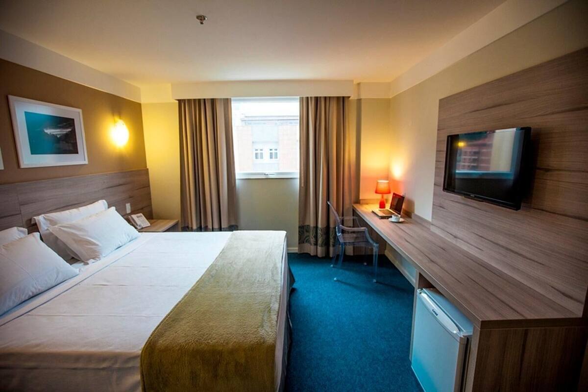Habitación de hotel con cama, escritorio, silla, televisor y cortinas.