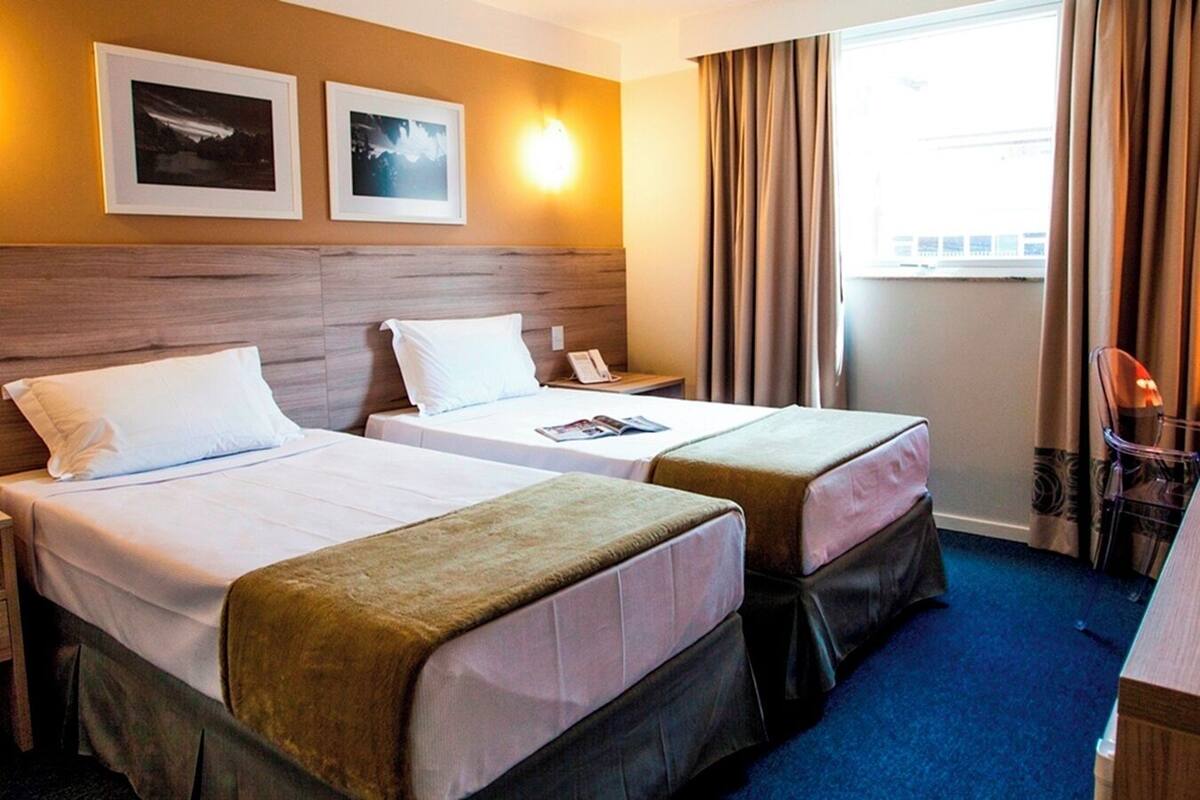 Habitación de hotel con dos camas, cabecera de madera, mesita de noche, una silla y una ventana con cortinas.