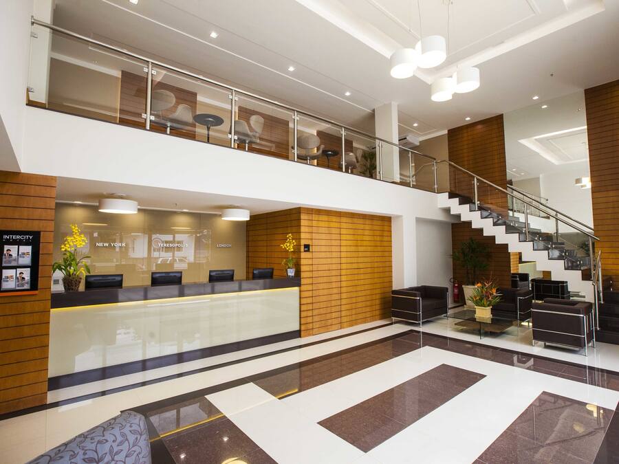 A modern hotel lobby with a reception desk, seating area, staircase, and a glass railing on the upper level.