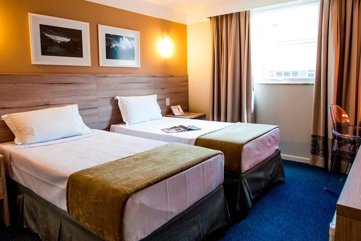 Habitación de hotel con dos camas, cabecero de madera, mesita de noche, una silla y ventana con cortinas.