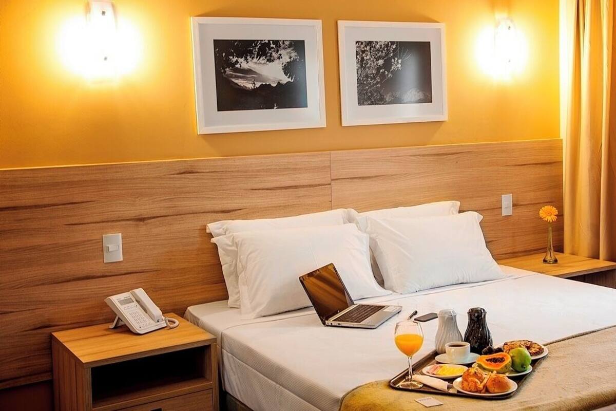 Habitación de hotel con cabecera de madera, cama con ropa blanca, mesita de noche con teléfono y ordenador portátil, y bandeja con artículos para el desayuno.