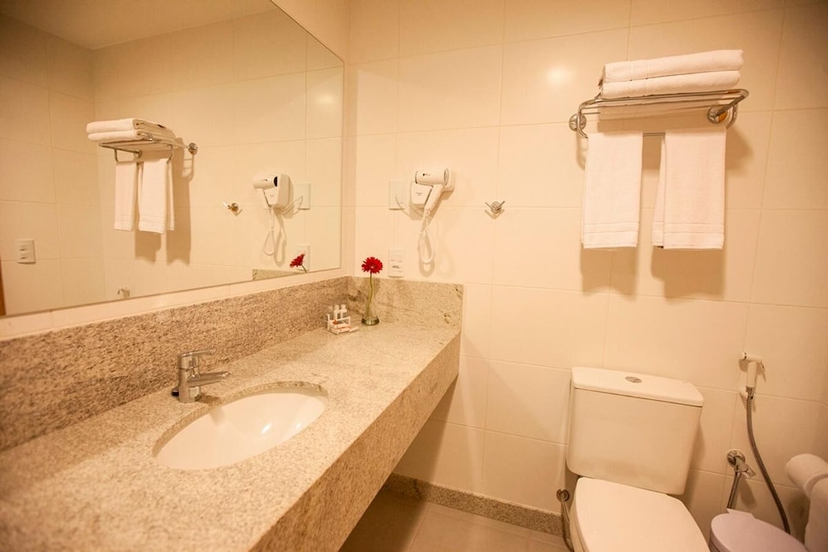 Casa de banho com lavatório de granito, sanita e dois secadores de cabelo fixados na parede.