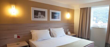 A hotel room with a large bed, two bedside tables, a nightstand, a lamp, a vase with a flower, two framed pictures on the wall, and a window with curtains.