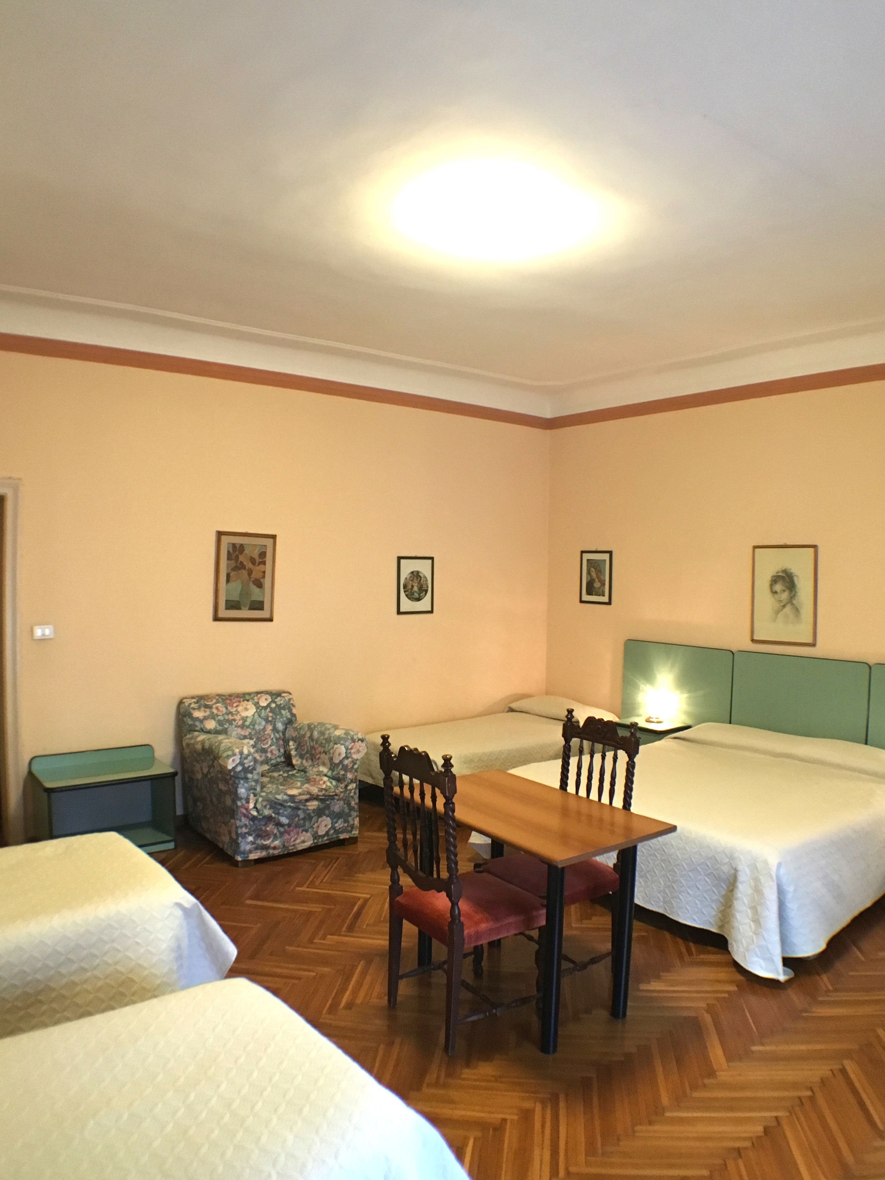 Une chambre avec deux lits, une table en bois, un fauteuil à motifs floraux et une petite table avec une lampe.