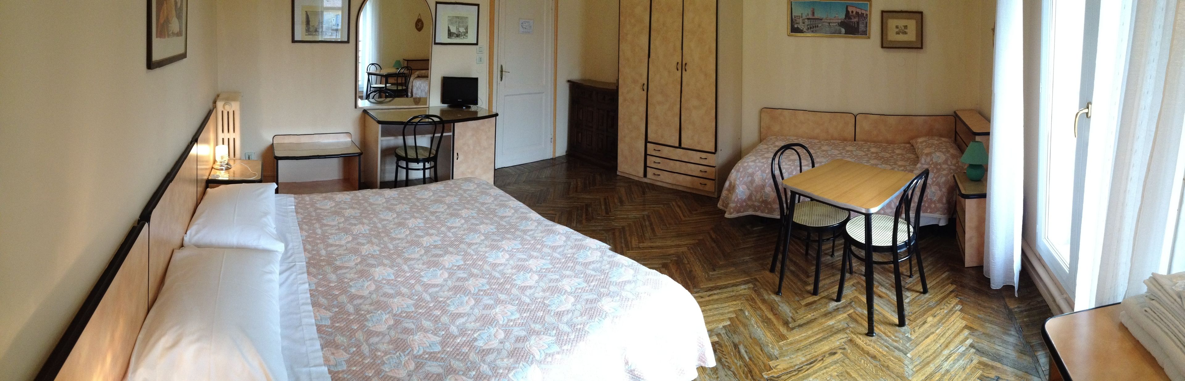 A bedroom with a bed, a desk, a chair, a wardrobe, and a small table with a lamp.
