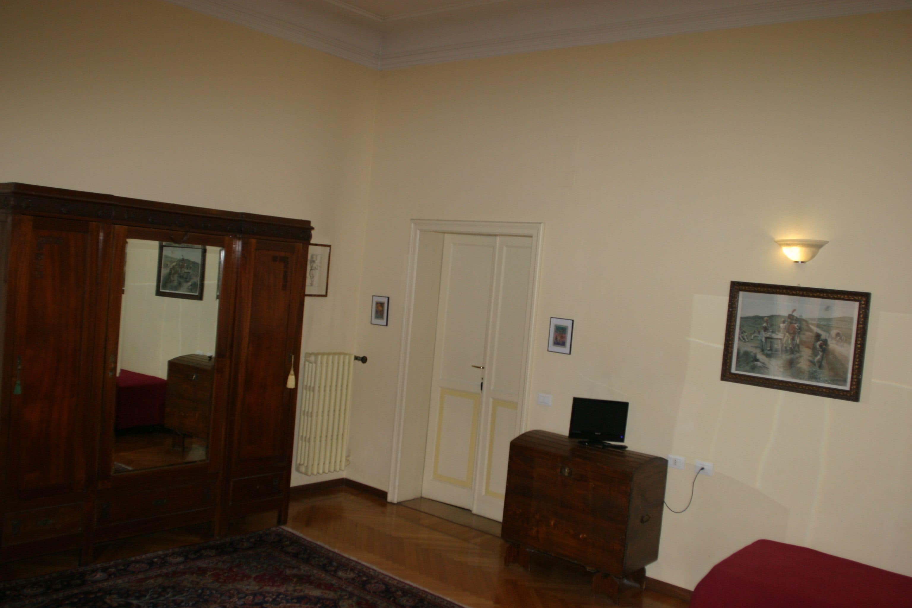 A room with a wooden wardrobe, a red sofa, a small wooden cabinet, and a framed picture on the wall.