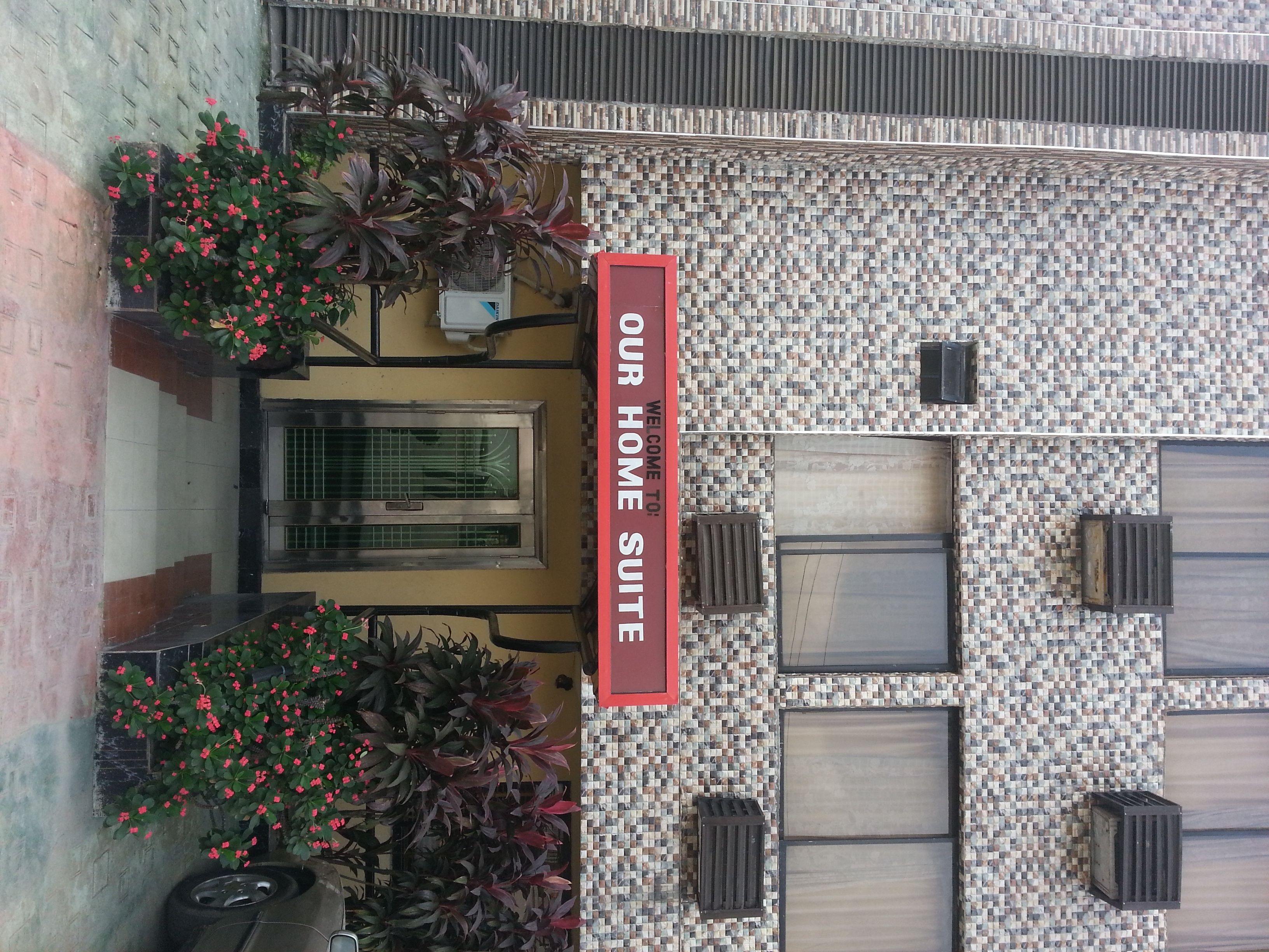 A building with a mosaic tile facade, a sign reading "OUR HOME SUITE," and potted plants with red flowers.