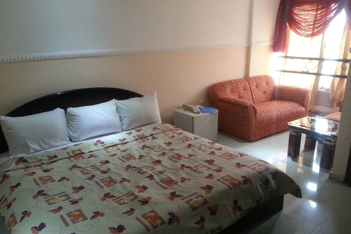 A hotel room with a bed, a small side table, a red armchair, and a glass table with a chair.