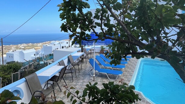A poolside area with blue lounge chairs and a pool, overlooking a coastal town with white buildings and the sea. - Stella Nomikou Apartments (Santorini)