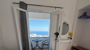 A hotel room with a bed, a small table, a chair, a balcony with a view, and a mirror. - Stella Nomikou Apartments (Santorini)