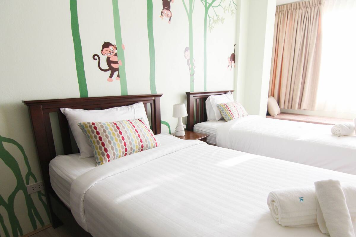 A hotel room with two beds, each with white bedding and a colorful pillow. There is a nightstand with a lamp and a window with curtains.