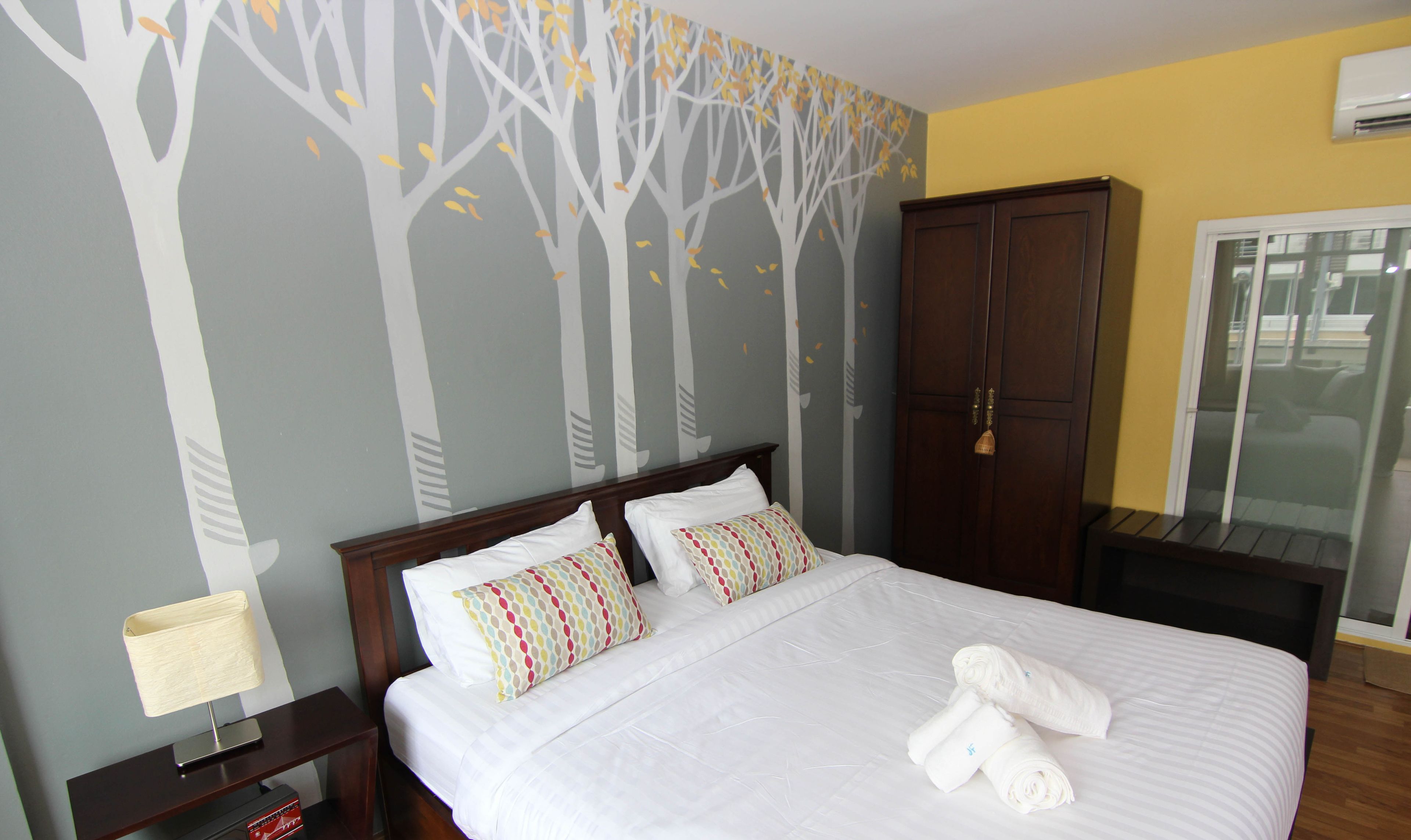 Una cama bien hecha con sábanas blancas y almohadas coloridas, una mesita de noche de madera con una lámpara, un armario y una ventana con vistas al exterior.