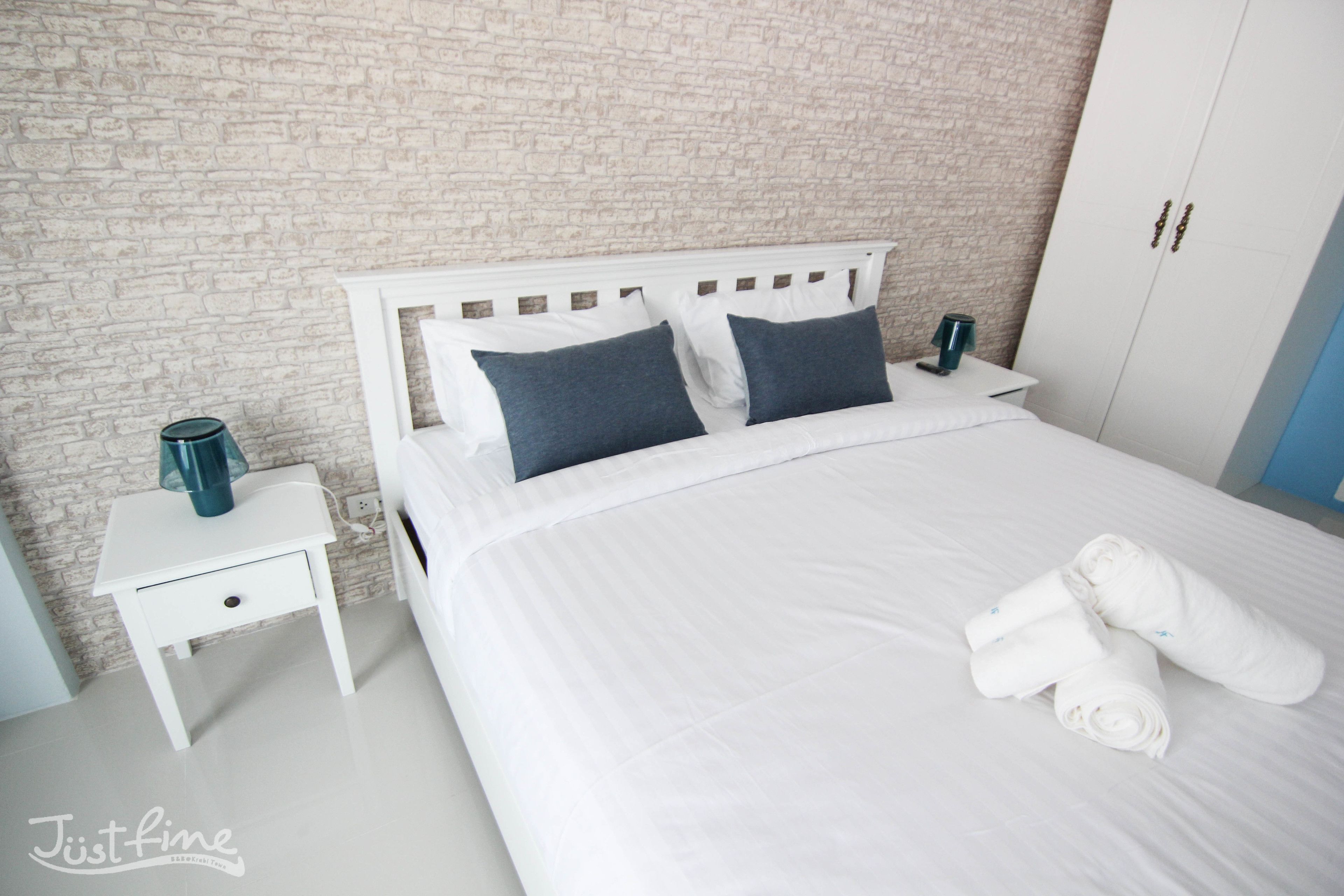 A neatly made bed with white bedding and two dark pillows, a white nightstand with a lamp, and a wardrobe with two doors.