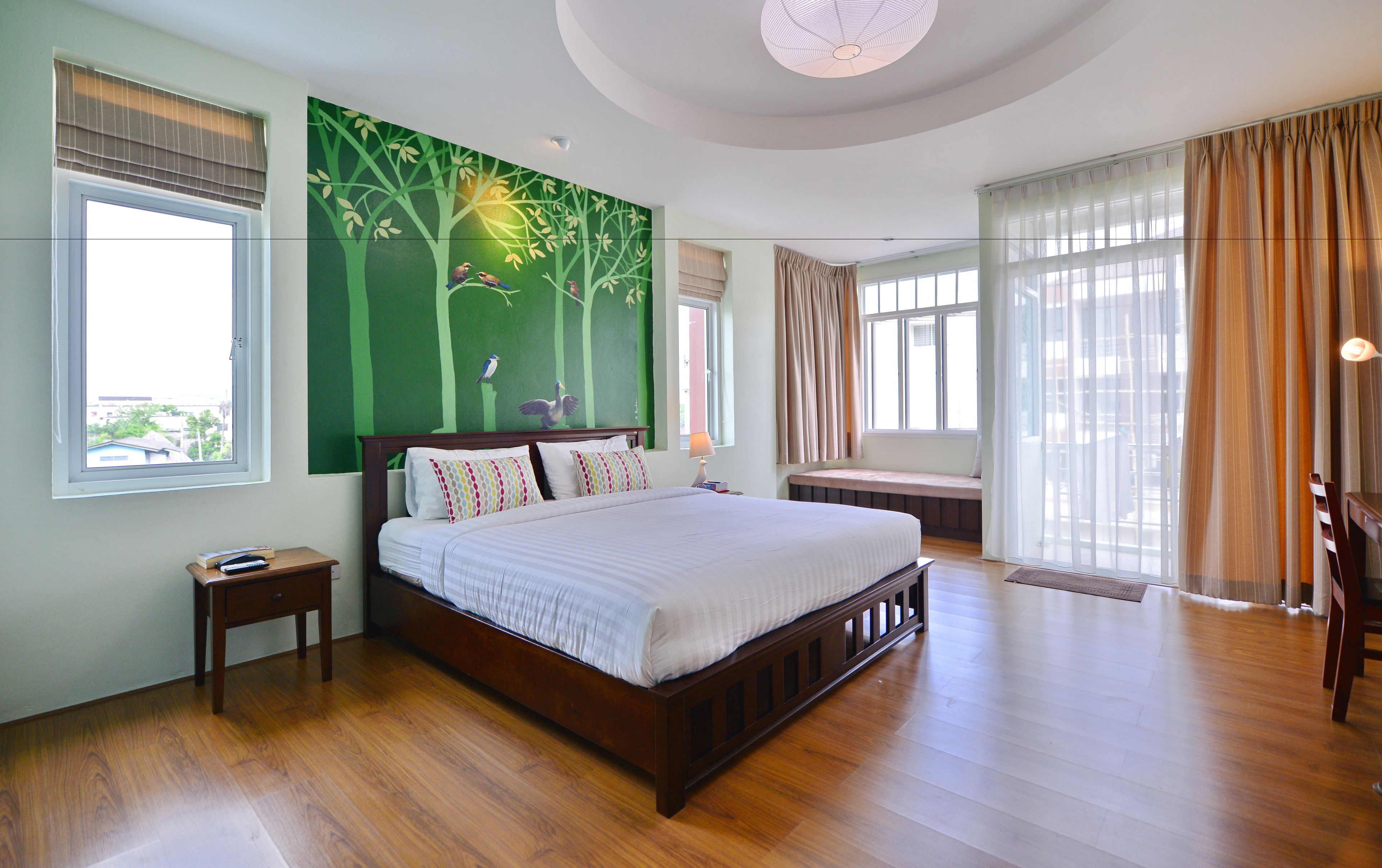 Une chambre spacieuse avec un grand lit, un plancher en bois, un mur décoré d’une fresque verte et de nombreuses fenêtres.