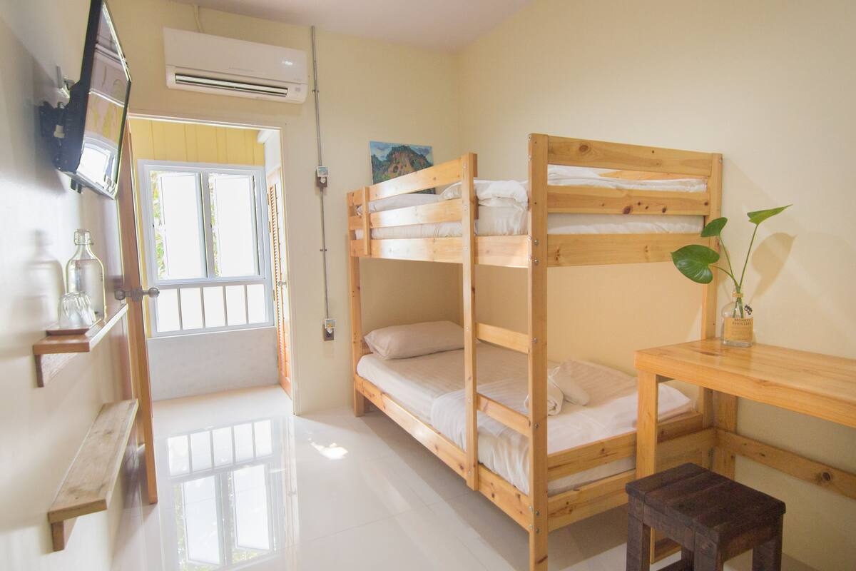 A bunk bed room with a wooden desk and bench, a TV mounted on the wall, and a window with curtains.