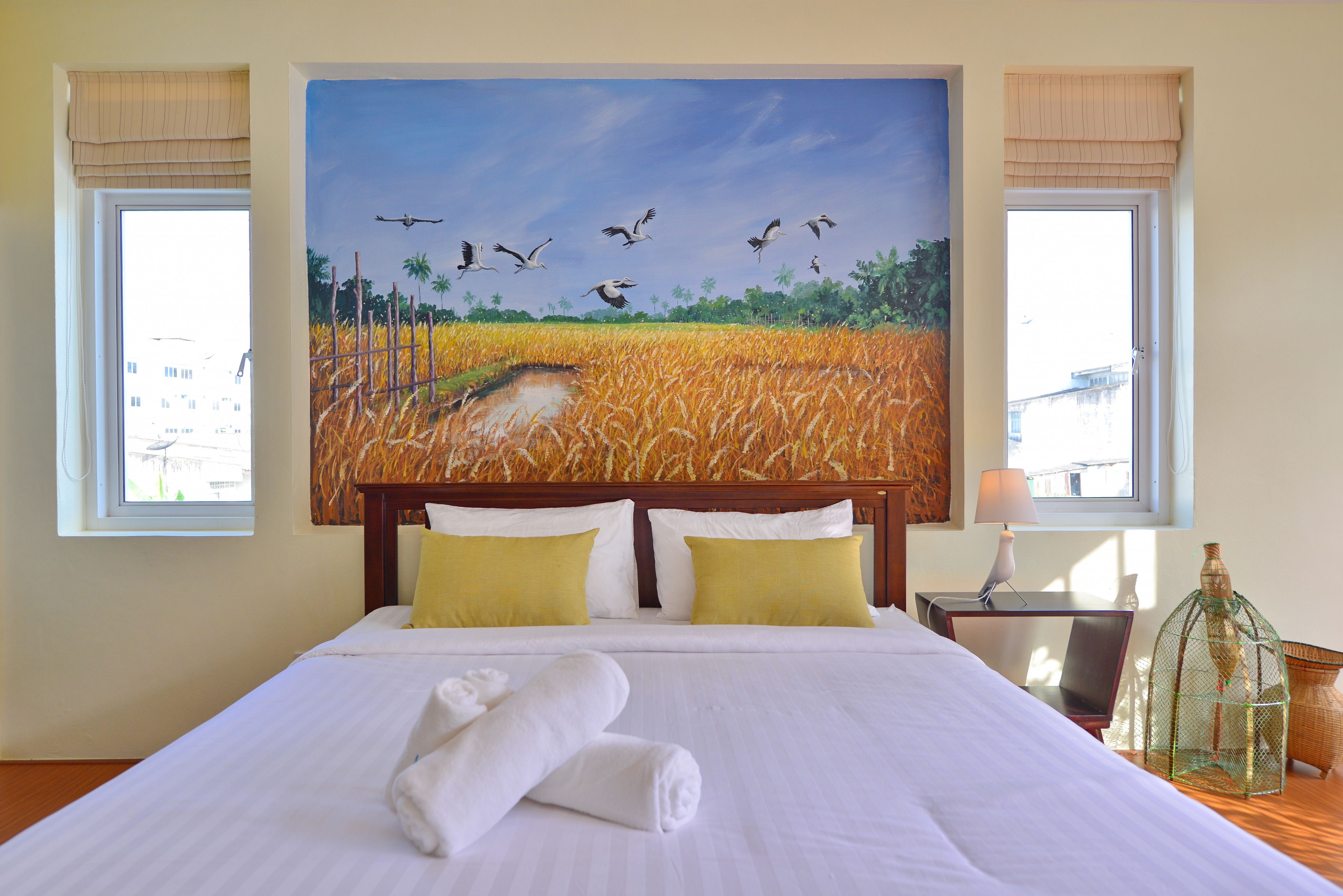 A hotel room with a large bed, two pillows, a lamp, a birdcage, and a painting of birds flying over a field.