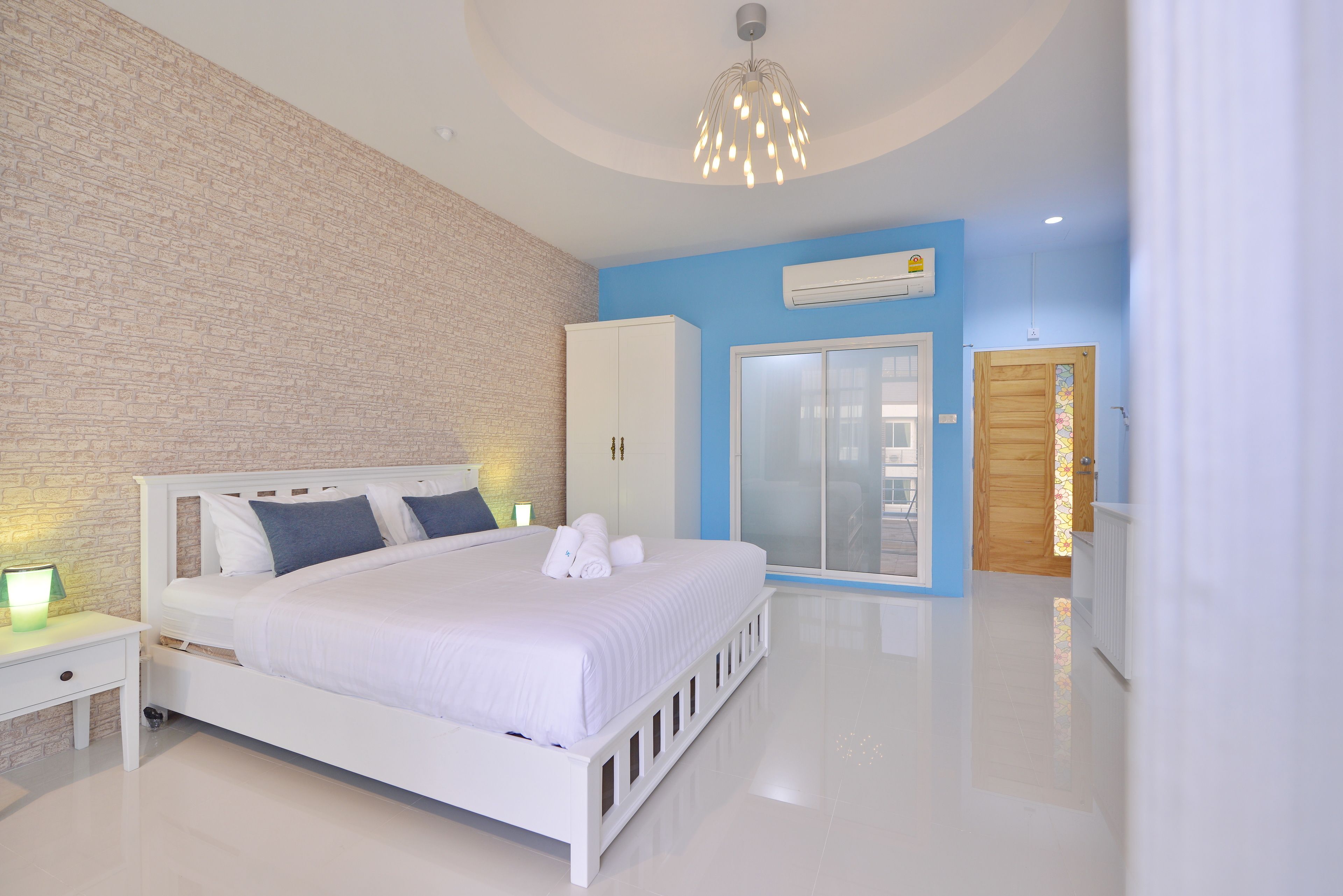 A modern bedroom with a white bed, a brick accent wall, a large window, and a wooden door.