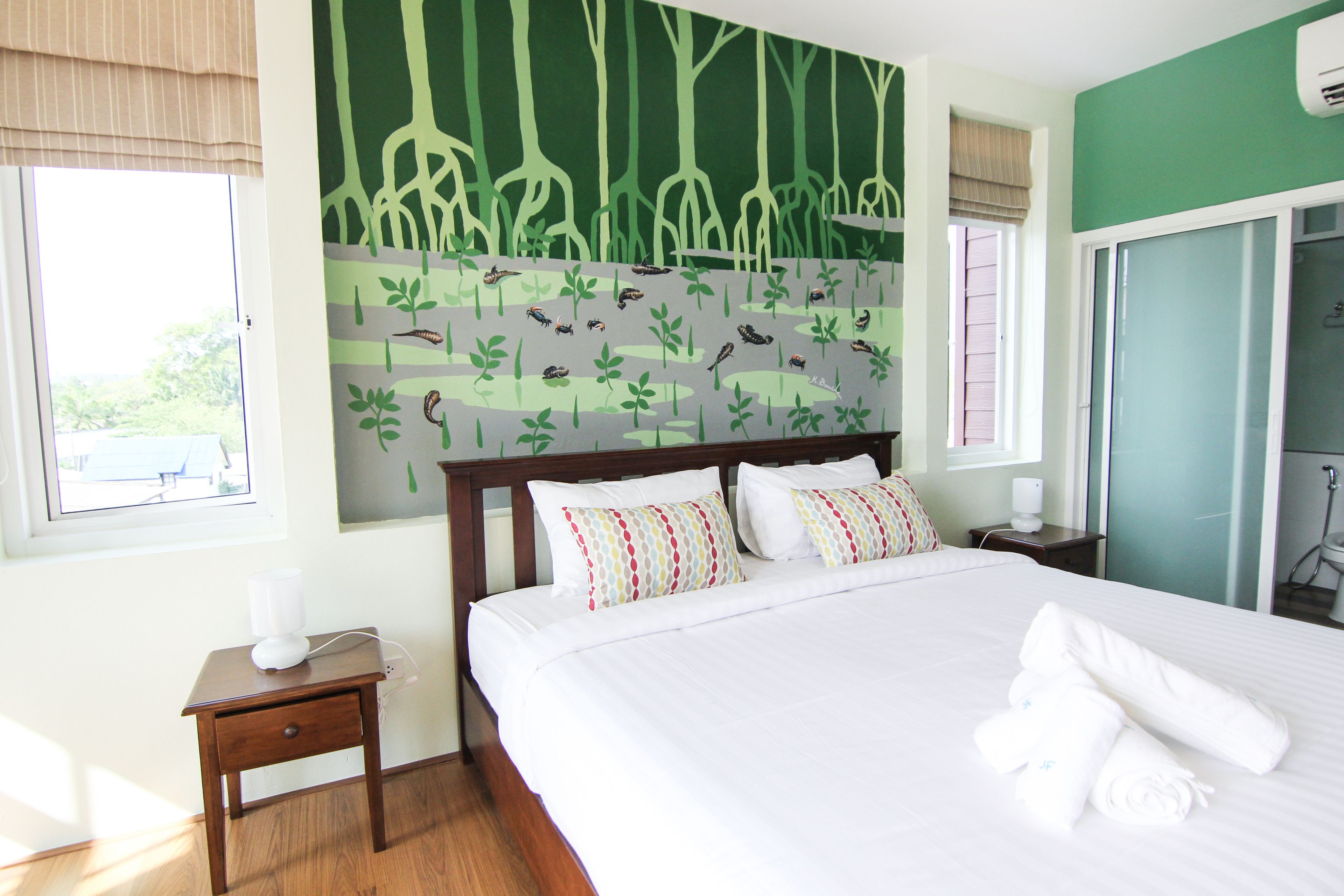 A neatly made bed with white linens and a set of towels, a bedside table with a lamp, and a wall mural of a forest scene.