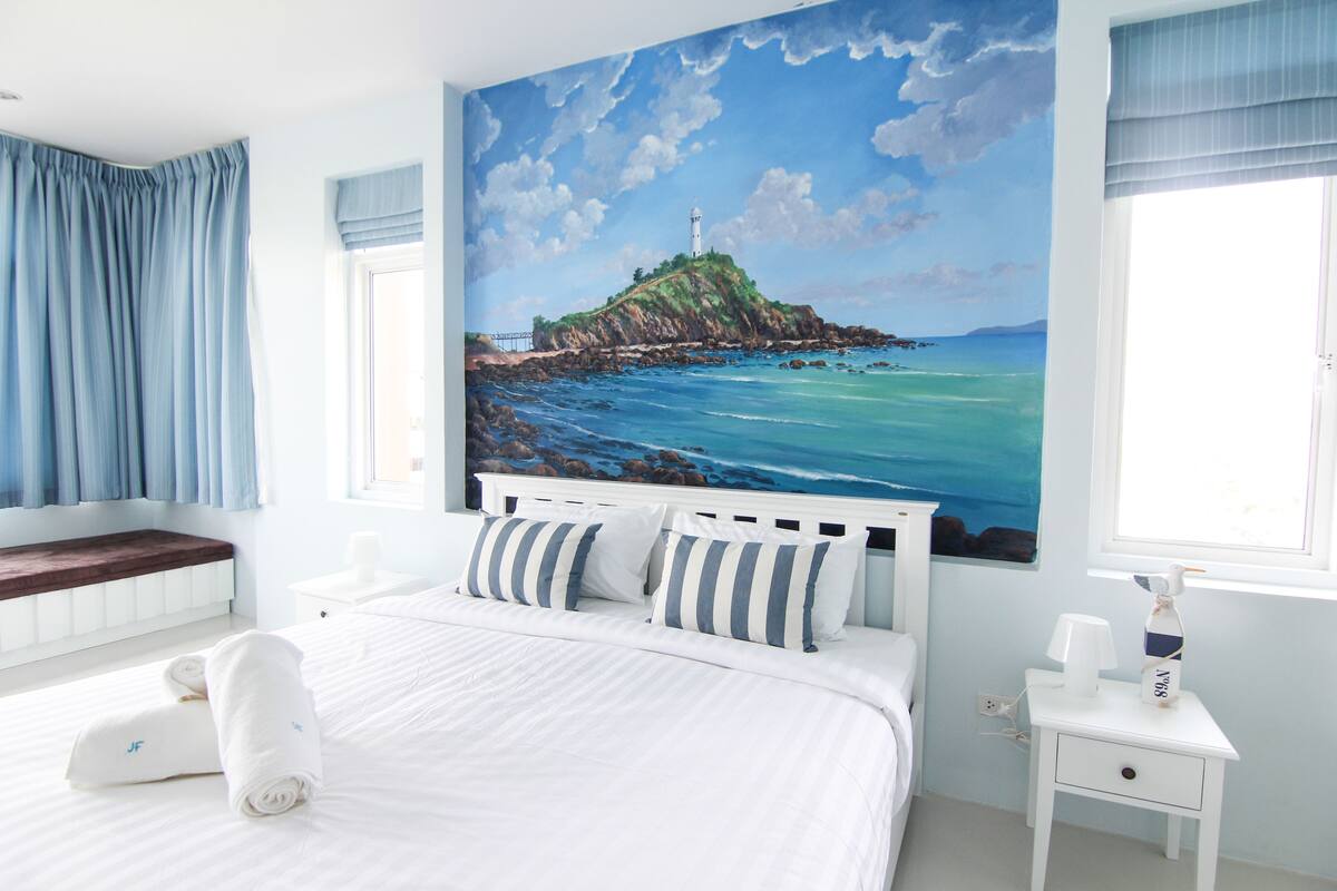 A bedroom with a large bed, a nightstand, a window with curtains, and a painting of a lighthouse on a cliff.