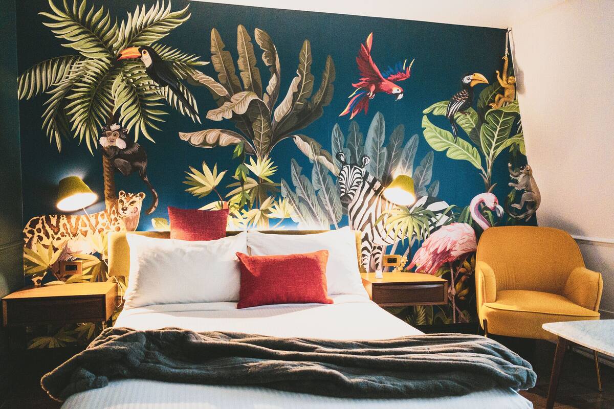 A bedroom with a bed, a nightstand, a lamp, a chair, and a wall mural.