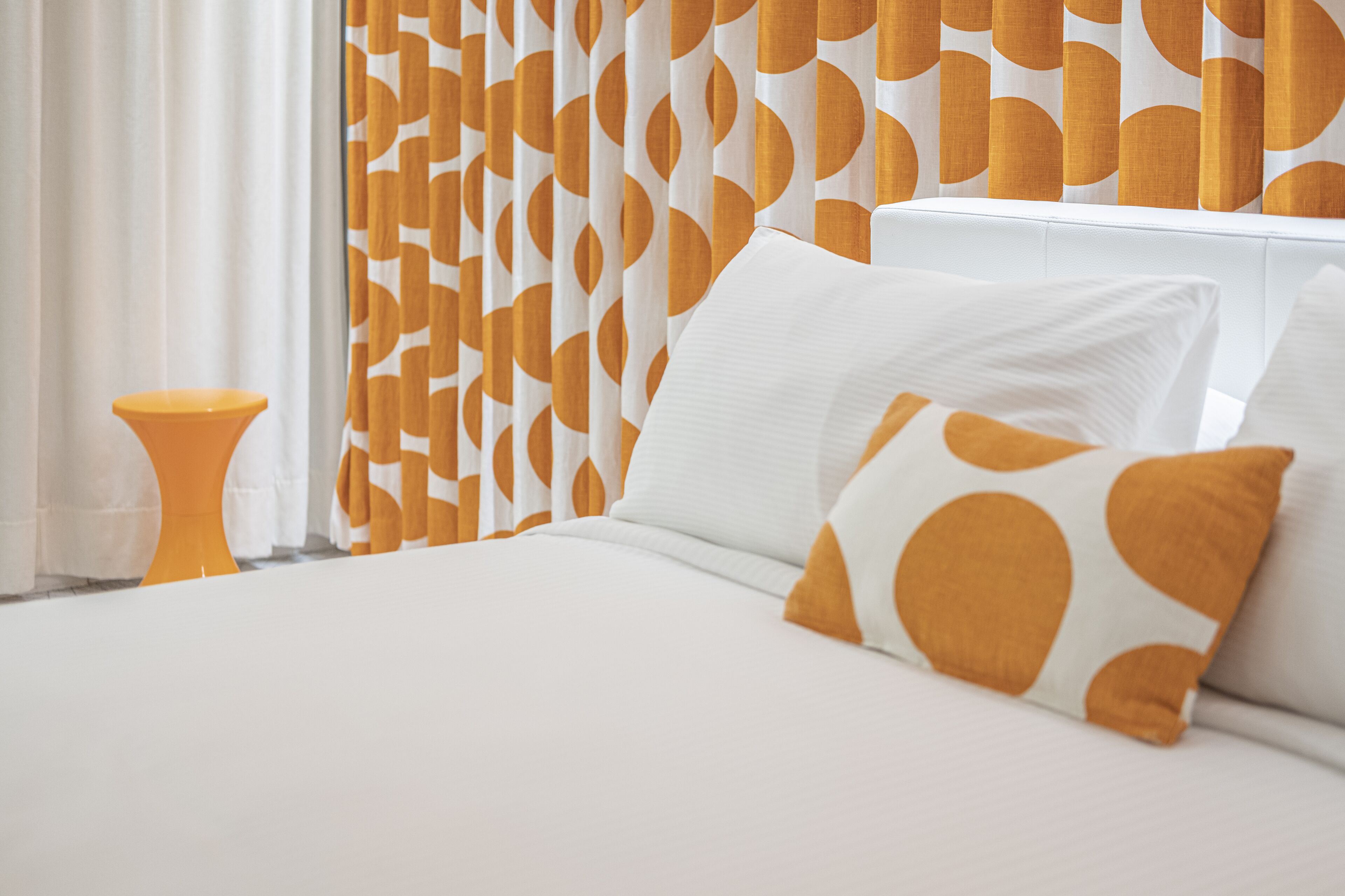 Una cama bien hecha con almohadas blancas y anaranjadas, un cabecero blanco y una cortina estampada al fondo.